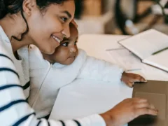 Smiling mother holding her baby while using a laptop at home, representing working moms balancing childcare and flexible family support.