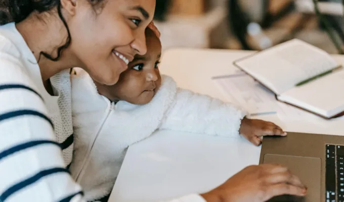 Smiling mother holding her baby while using a laptop at home, representing working moms balancing childcare and flexible family support.