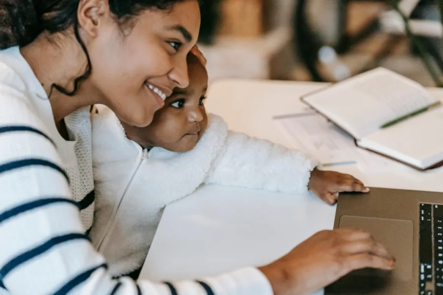 Smiling mother holding her baby while using a laptop at home, representing working moms balancing childcare and flexible family support.